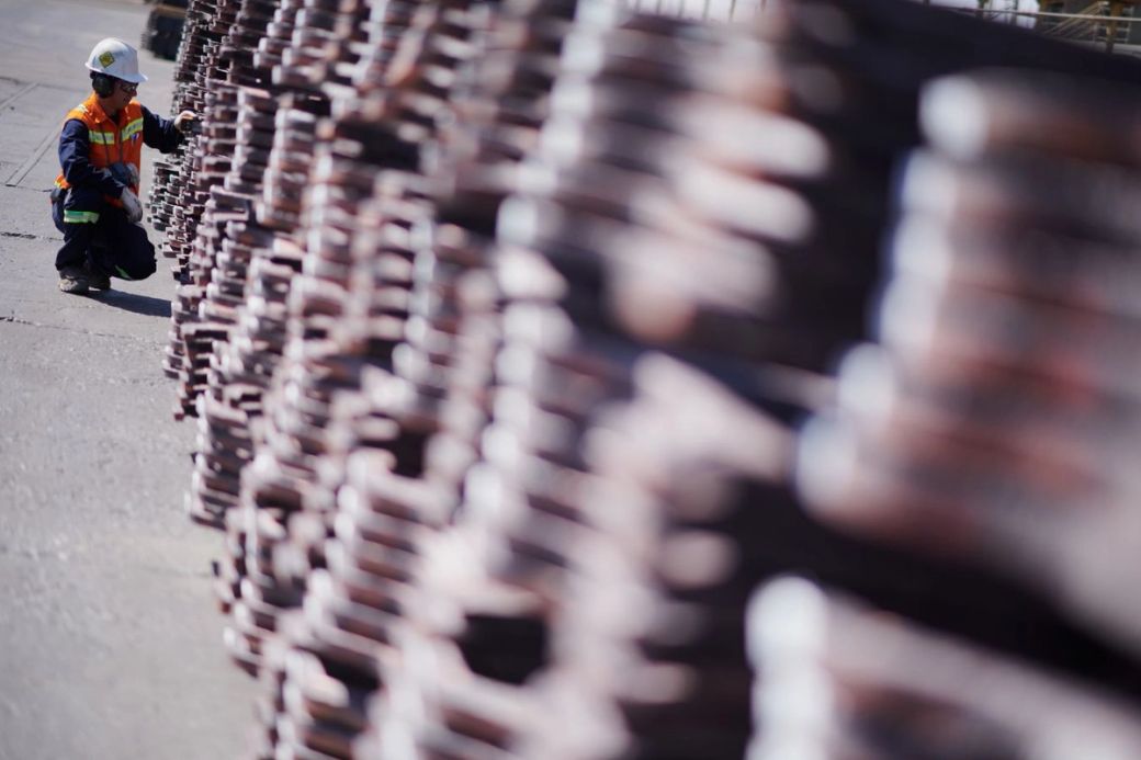 A construction worker in a safety vest kneels beside a stack of metal bars at a construction site. The focus is on the worker, while the metal bars create a blurred background.