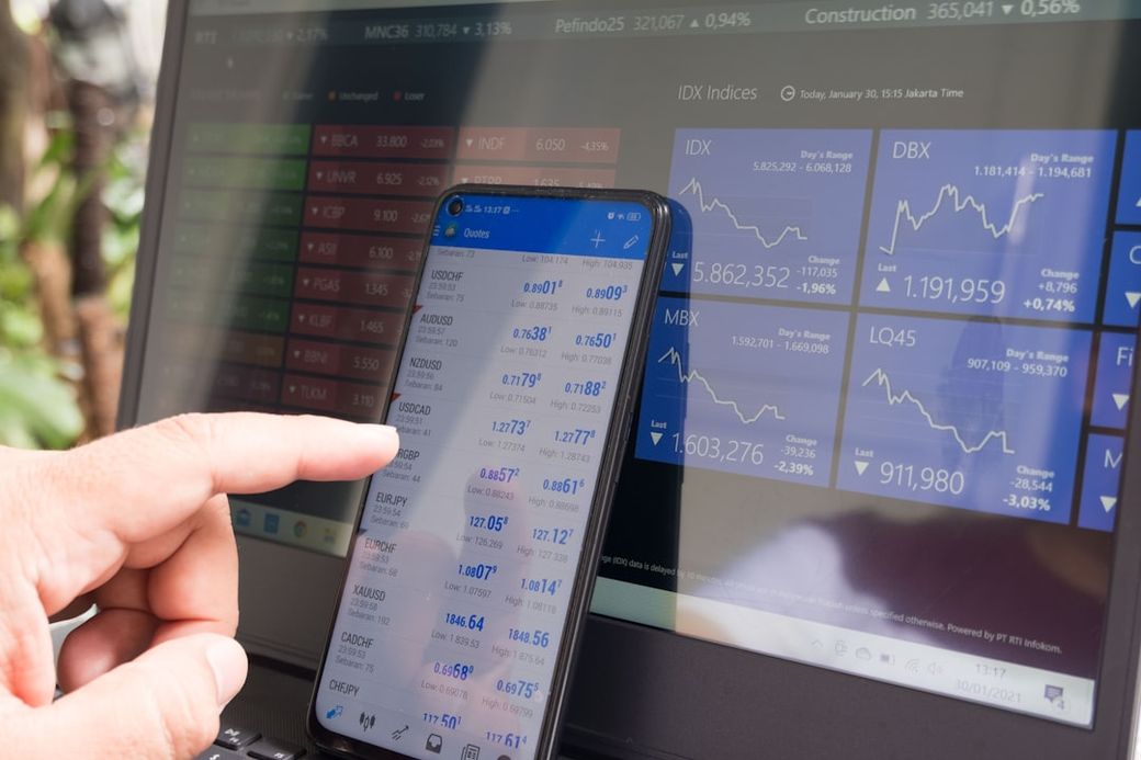 The image shows a close-up of a hand pointing at a smartphone displaying stock market data, next to a laptop with various financial indicators and charts on the screen. The background features a digital display of multiple stock indices and values. — Credit: Photo by Marga Santoso on Unsplash
