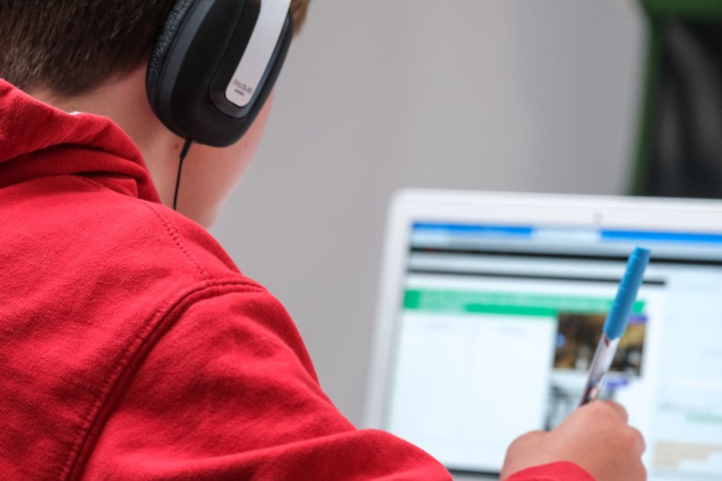 A young boy wearing headphones is focused on a computer screen while holding a blue pen. The setting appears to be a home or study environment, suggesting he is engaged in learning or digital activities. — Credit: Photo by Compare Fibre on Unsplash