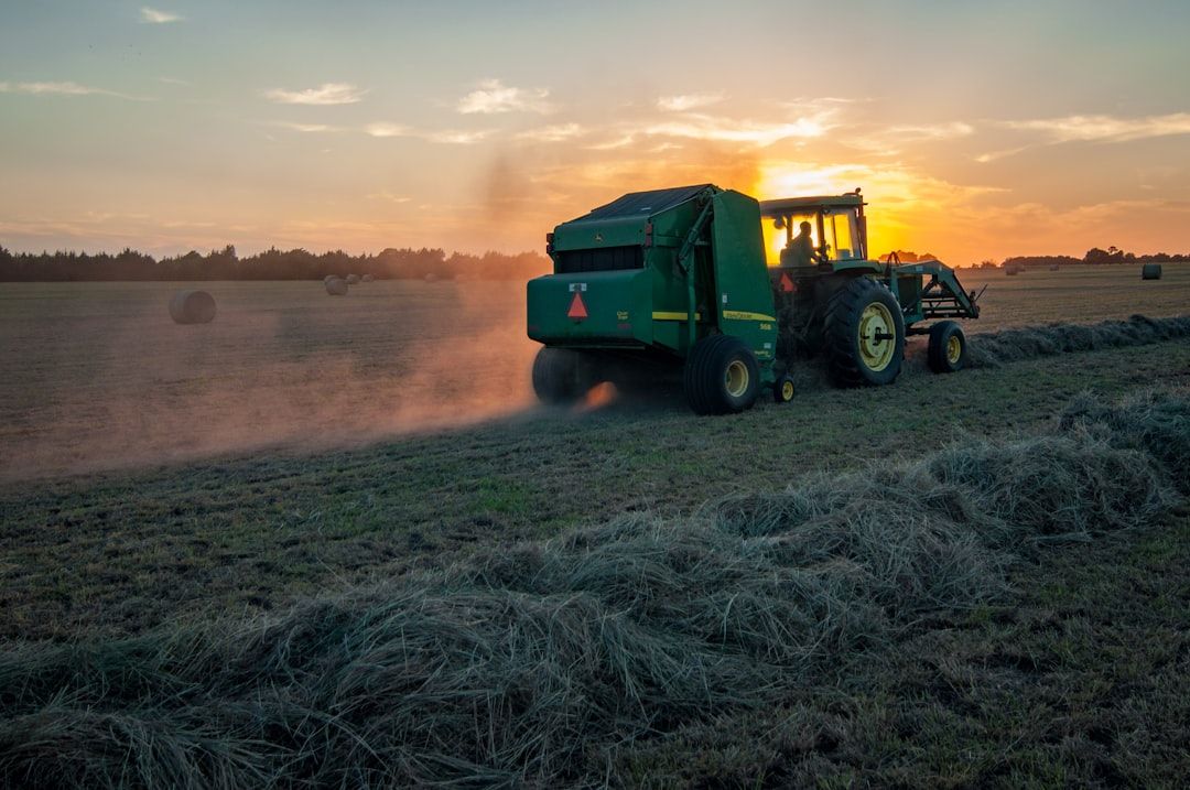 A tractor is seen working in a field during sunset, creating dust as it moves along a row of hay. The warm colors of the sunset illuminate the scene, highlighting the agricultural activity.