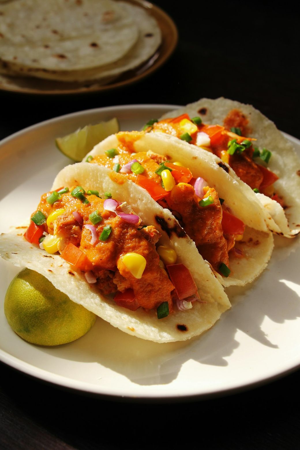 The image features three tacos filled with orange-hued fish, garnished with diced tomatoes, corn, and chopped onions, served on a plate alongside a lime wedge. A stack of tortillas is visible in the background. — Credit: Photo by montatip lilitsanong on Unsplash