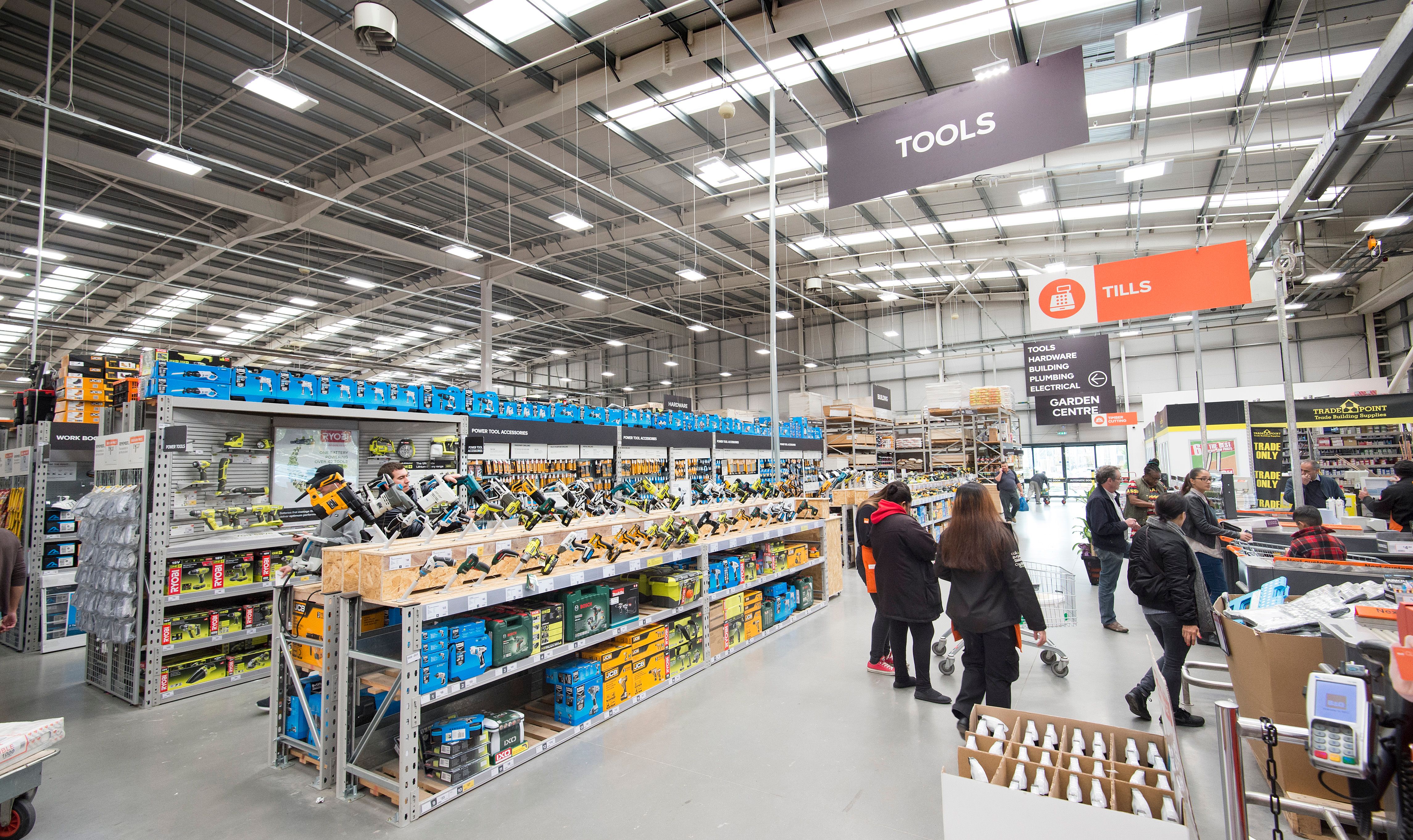The image shows the interior of a large retail store, B&Q, specifically a hardware or home improvement store. The aisles are well-stocked with various tools and equipment, and customers can be seen shopping.