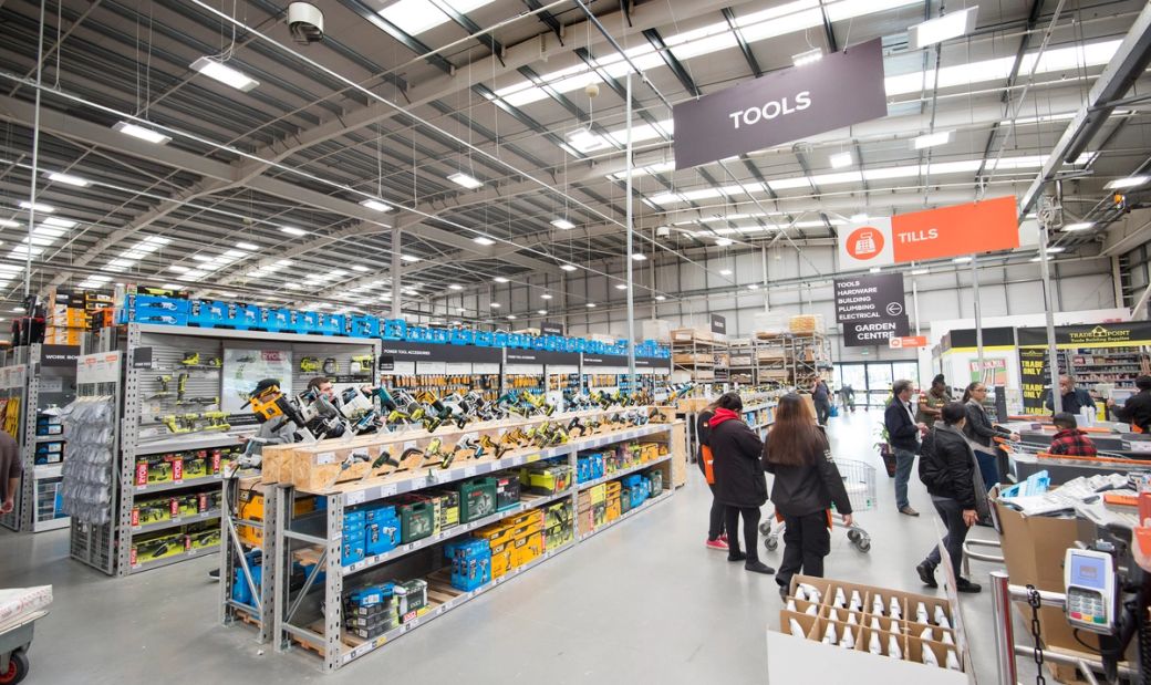 The image shows the interior of a large retail store, B&Q, specifically a hardware or home improvement store. The aisles are well-stocked with various tools and equipment, and customers can be seen shopping.