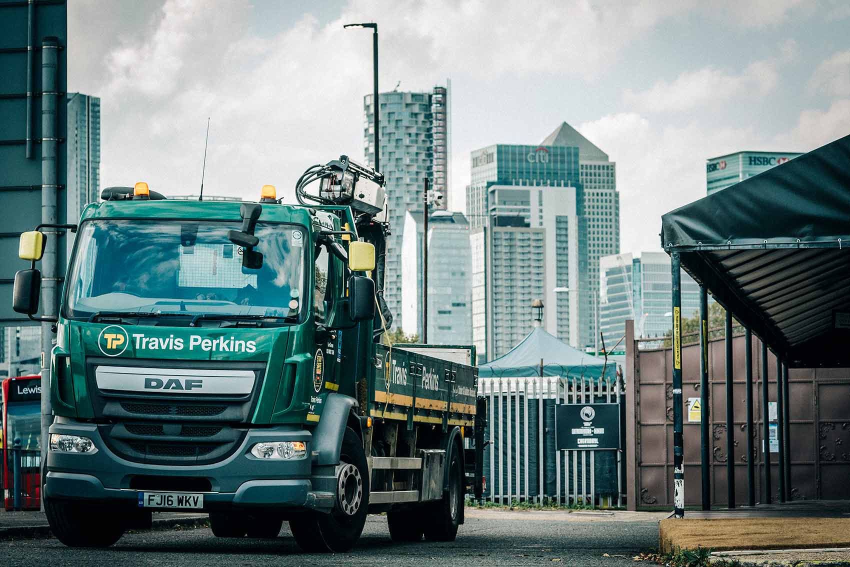 A green DAF truck from Travis Perkins is seen approaching a construction site with a backdrop of modern skyscrapers. The truck appears to be part of a logistical operation in an urban environment.