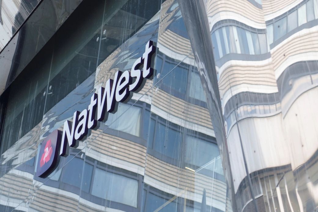 The image shows the exterior of a modern building featuring the NatWest bank logo prominently displayed. The reflective surface of the building creates an interesting visual effect with surrounding architecture. — Credit: Dominic Lipinski/NatWest Group