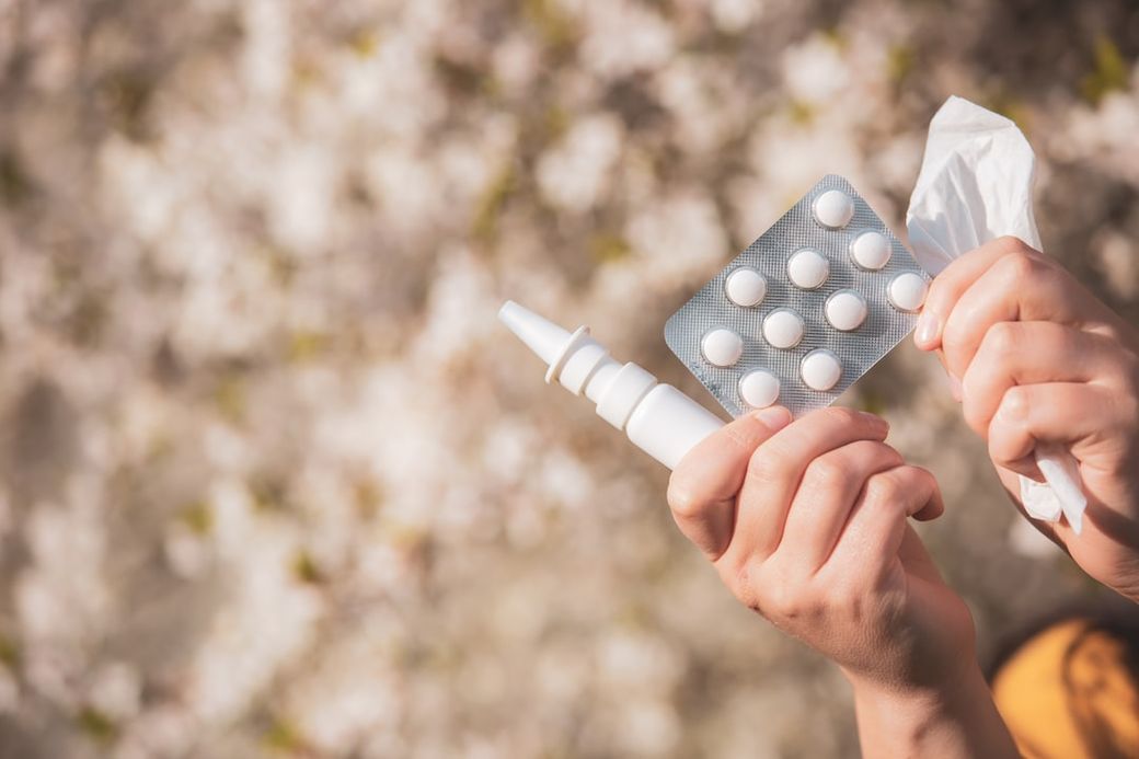 The image features a person holding a nasal spray and a blister pack of white tablets, suggesting a focus on medication and health. The background consists of soft, blurred floral elements, adding a calming aesthetic. — Credit: Photo by Simon Kadula on Unsplash