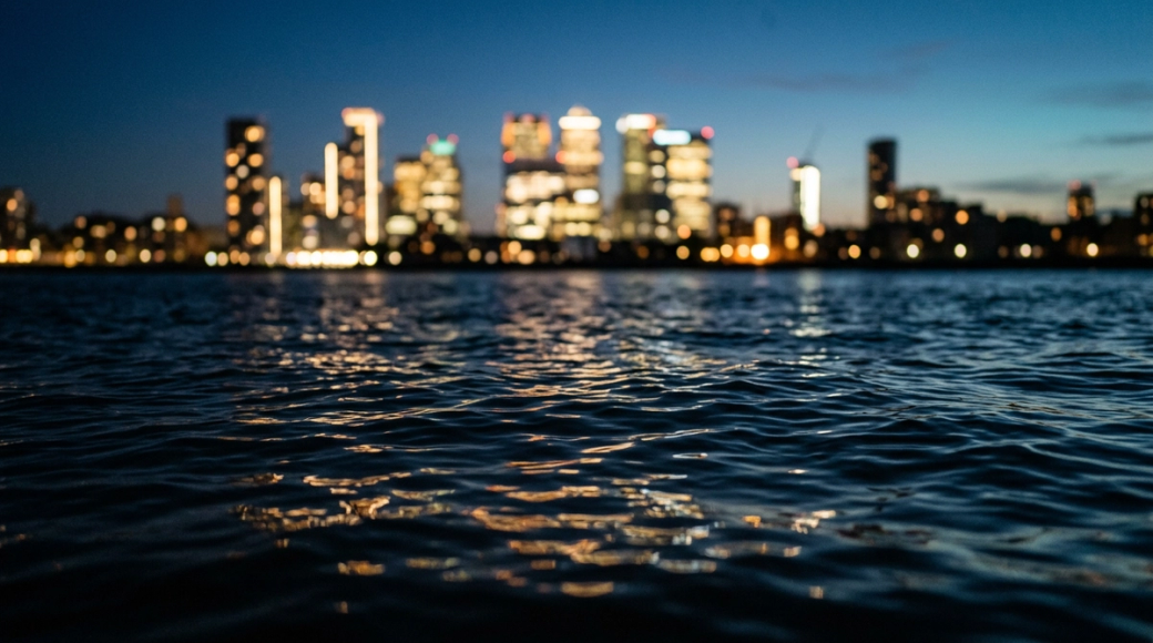 The image captures a serene view of the Thames at blue hour, with the river's surface dominating the foreground. The Canary Wharf skyline appears softly blurred in the background, creating a contrast between the detailed ripples of the water and the abstract luminosity of the buildings, highlighting themes of wealth and infrastructure.