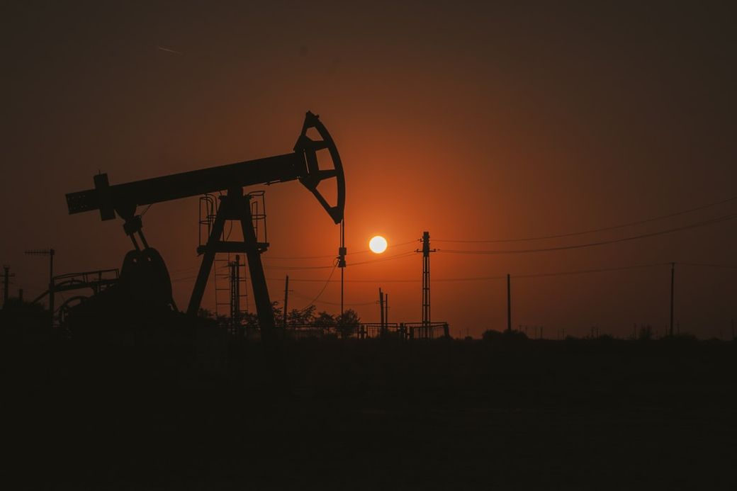 The image captures a silhouetted oil pump jack against a sunset backdrop, with the sun setting on the horizon. Power lines and a desolate landscape add to the industrial aesthetic. — Credit: Photo by Mihai 👑 on Unsplash