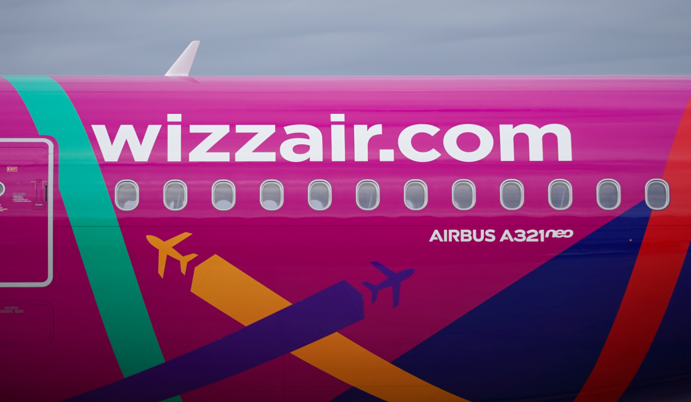The image showcases the side of an Airbus A321neo aircraft belonging to Wizz Air, featuring a vibrant pink exterior with colorful geometric patterns. The airline's website is prominently displayed.
