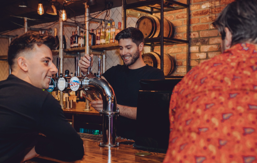 A bartender is serving drinks at a lively bar, with patrons engaging in conversation. The setting features wooden barrels and a well-stocked shelf of alcoholic beverages in the background.