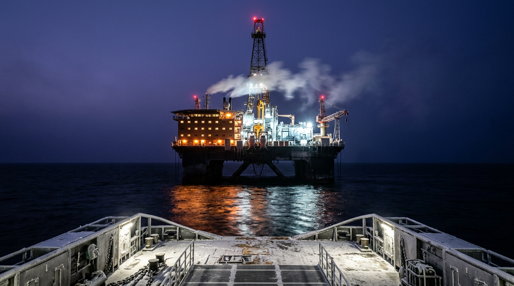 The image captures a semi-submersible rig in the Barents Sea during polar twilight, illuminated by its own ambient lighting. The rig's accommodation block glows with warm lights, while the drill floor is bathed in cold white floodlight, with red warning lights on the derrick. The deck of a supply vessel is visible in the foreground, showcasing frost on equipment against a backdrop of dark, reflective water.