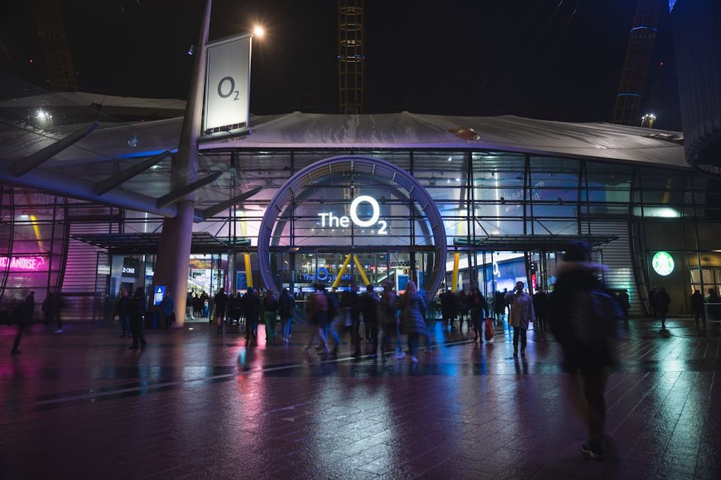 The image showcases the exterior of The O2, a prominent entertainment venue in London, illuminated with colorful lighting at night. People are seen milling around outside the entrance, creating a lively atmosphere. — Credit: Photo by Sandra Tan on Unsplash