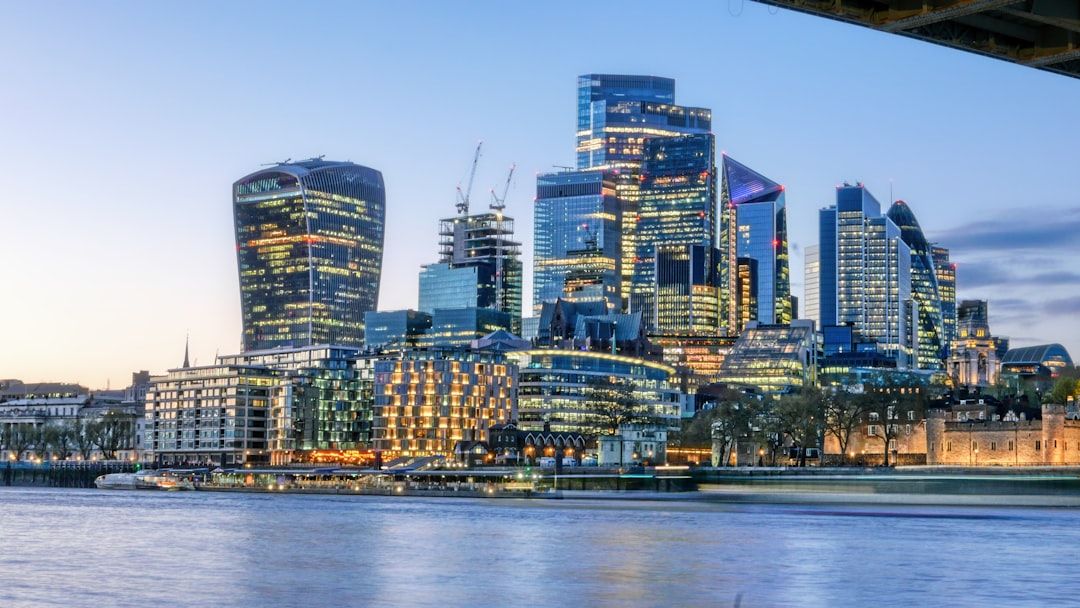 The image captures the skyline of London, showcasing a collection of modern skyscrapers along the River Thames during twilight. The buildings are illuminated, reflecting a vibrant city atmosphere.