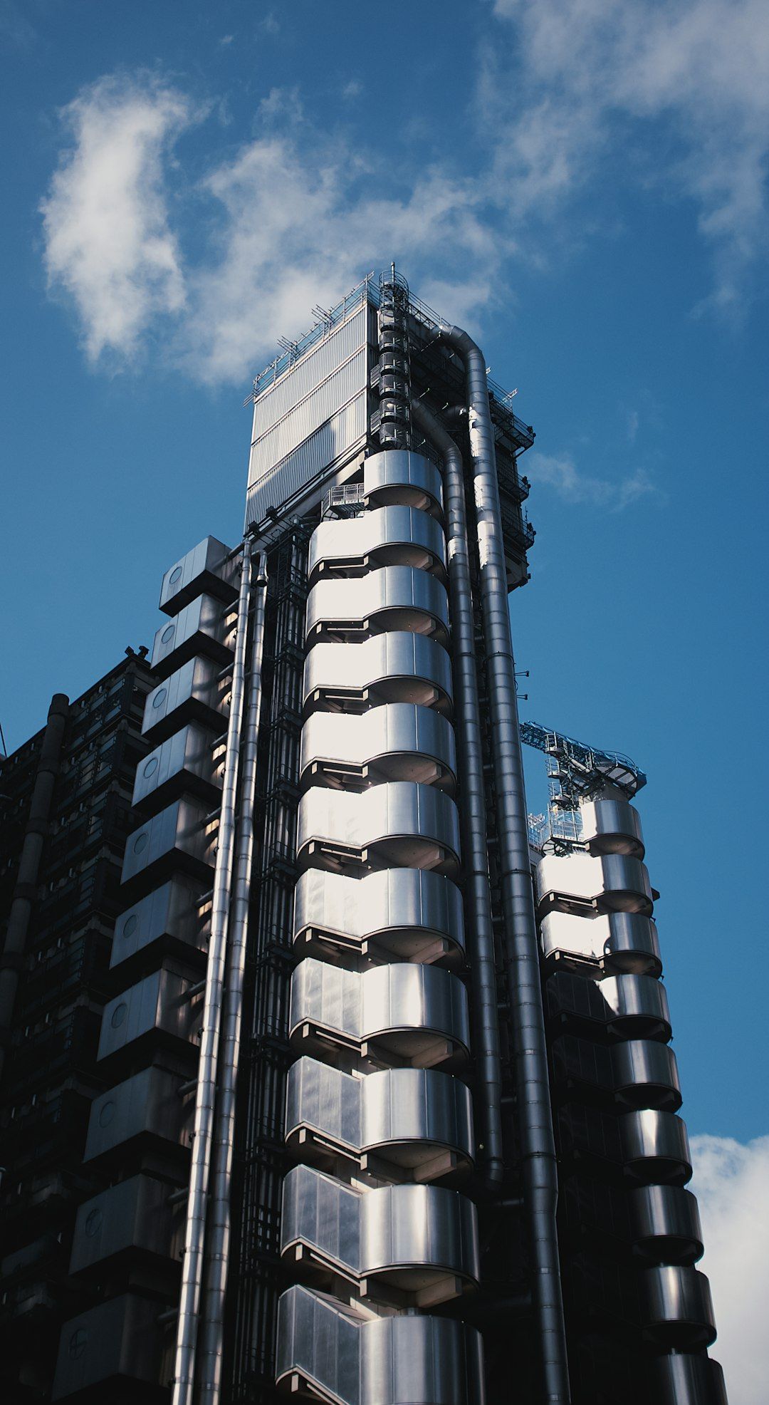 The image features a modern architectural structure characterized by metallic surfaces and exposed piping, set against a clear blue sky. The building's design features a unique vertical alignment with cylindrical elements visible on its facade.