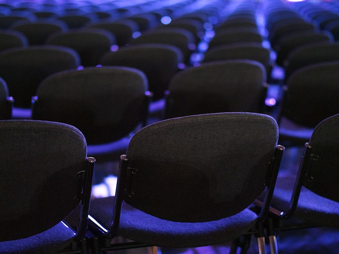 The image captures a close-up view of rows of empty black chairs arranged in a theater or conference setting. The chairs are focused in the foreground with a blurred background, creating a sense of depth.