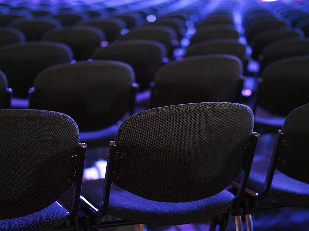 The image captures a close-up view of rows of empty black chairs arranged in a theater or conference setting. The chairs are focused in the foreground with a blurred background, creating a sense of depth. — Credit: Photo by Andrei Stratu on Unsplash