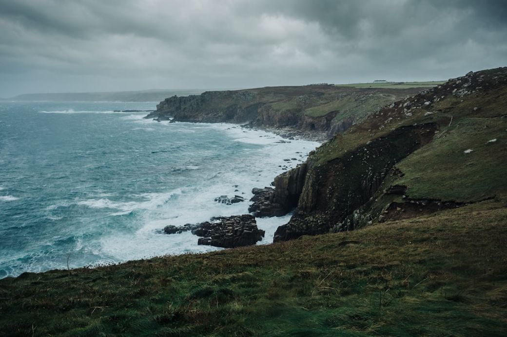A rugged coastline features dramatic cliffs and crashing waves under a cloudy sky. The landscape showcases the natural beauty and wildness of the coastal environment. — Credit: Photo by Andy Holmes on Unsplash