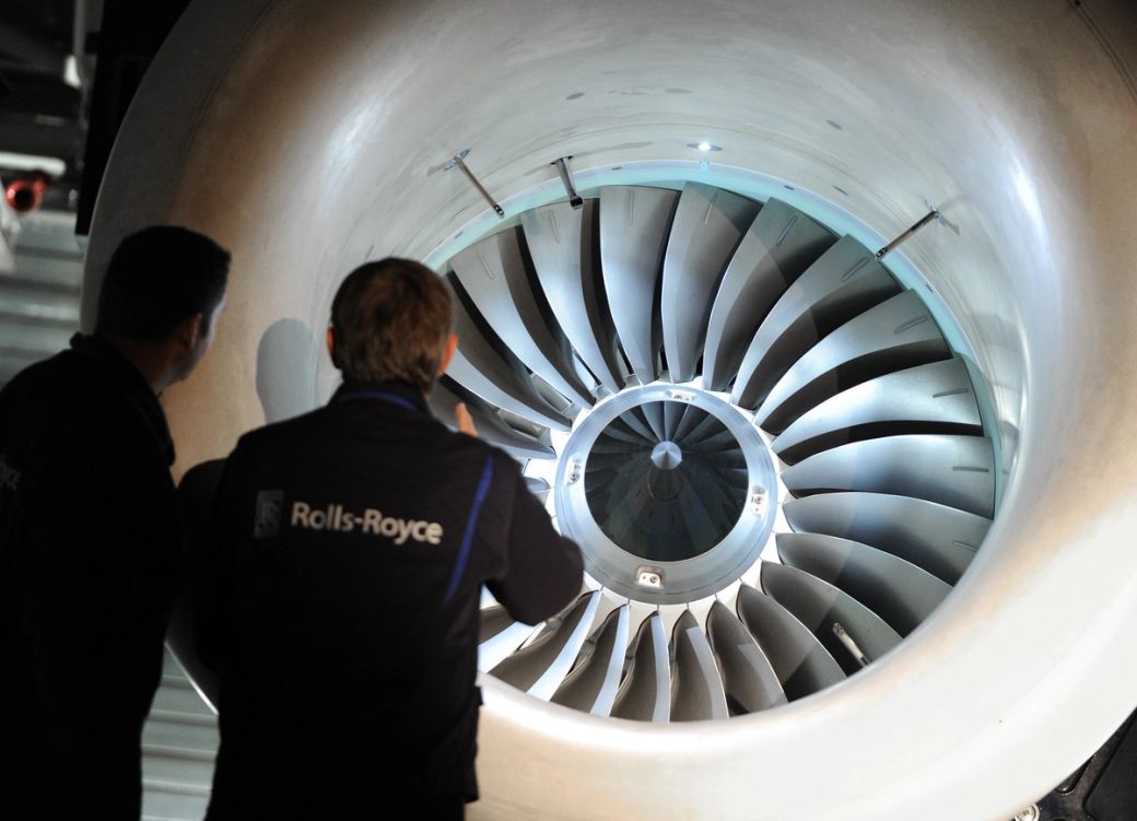 Two technicians examine a large jet engine turbine, highlighting its intricate design and engineering. The scene depicts a close-up view of the engine, emphasizing the technology involved in aircraft manufacturing.