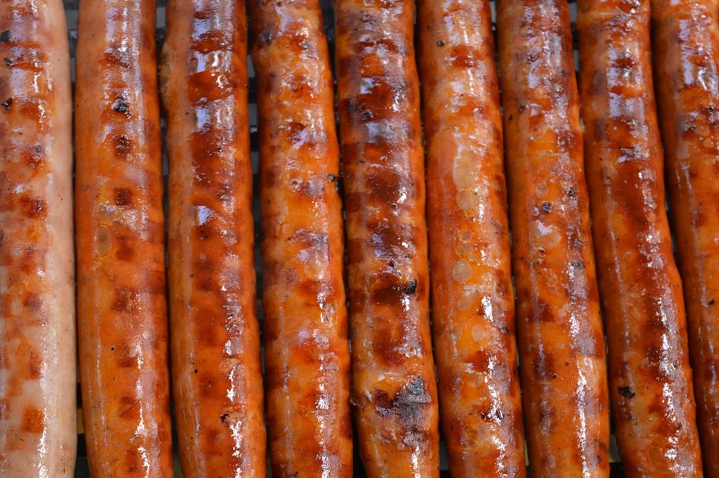 The image features multiple grilled sausages arranged in a row, showcasing their golden-brown, charred exterior. The sausages are neatly lined up on a grill surface, suggesting a cooking or barbecue setting. — Credit: Photo by Marko Anastasijevic on Unsplash