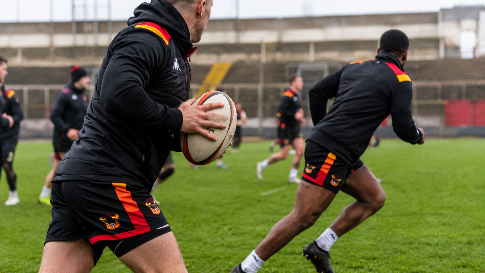 The image shows a rugby practice session with players actively training on a grassy pitch. One player is preparing to throw a rugby ball while another player runs in the background.