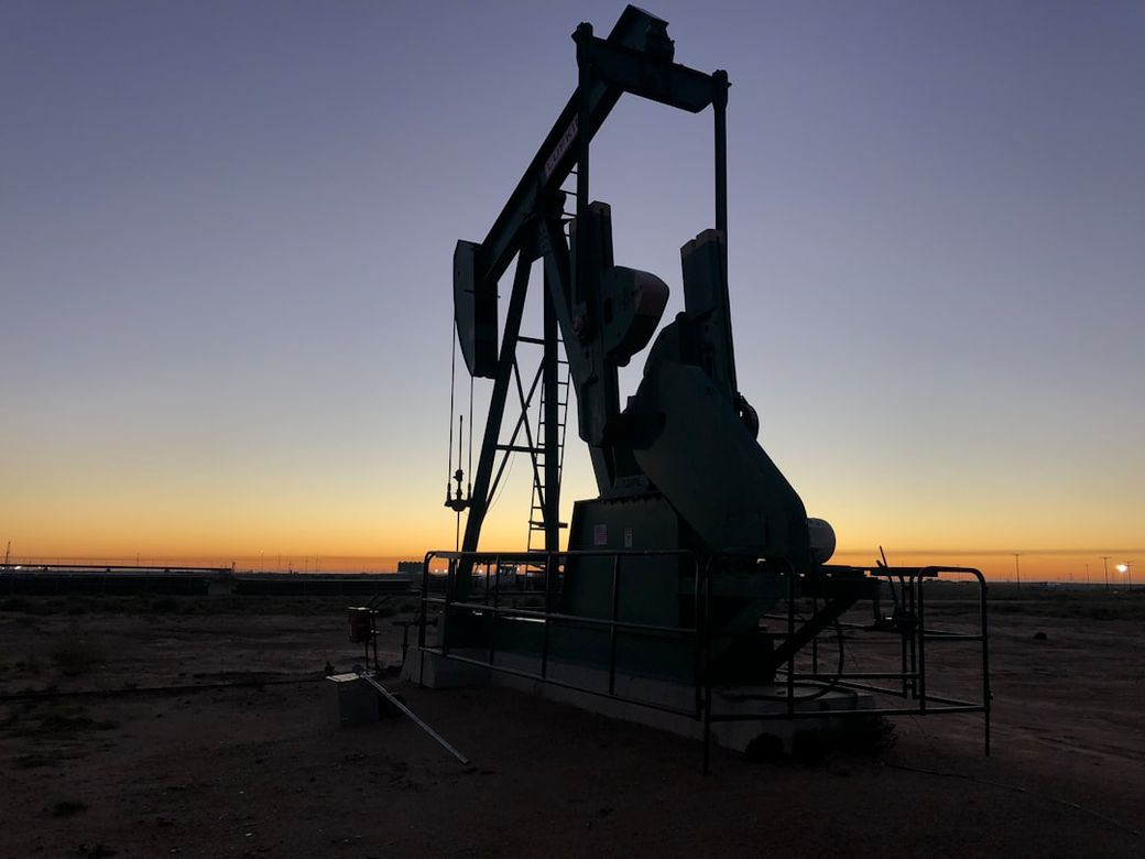 The image shows a silhouette of an oil pumpjack against a gradient sky during sunrise or sunset. The landscape appears arid, with a few structures visible in the background. — Credit: Photo by Delfino Barboza on Unsplash