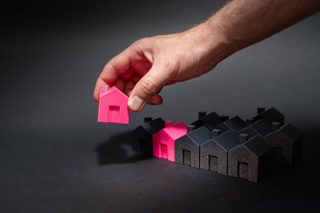 The image depicts a hand holding a small pink house while several black and dark gray houses are arranged in the background. The contrasting colors highlight the pink house, suggesting a focus on real estate or property selection.