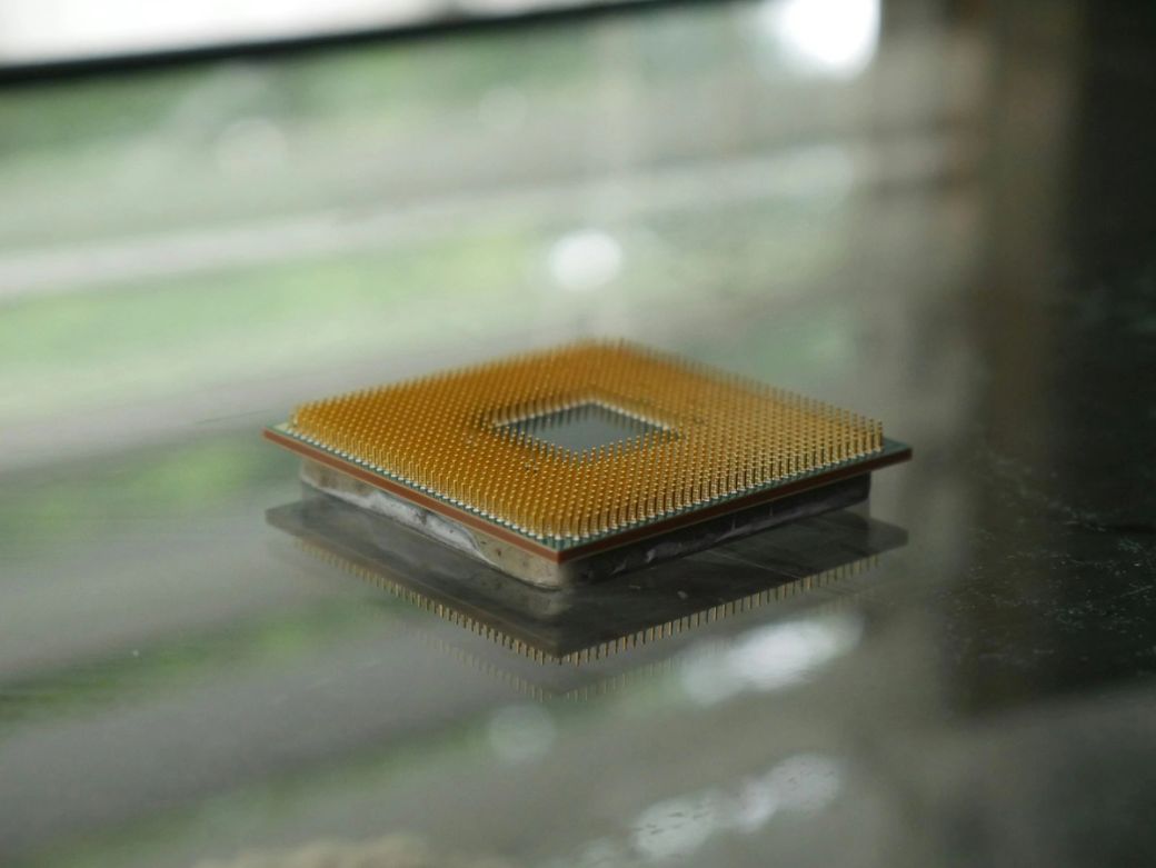 The image features a close-up view of a computer microprocessor resting on a reflective surface. The processor showcases a grid of gold pins and a square shape, highlighting its intricate design. — Credit: Photo by Devin Spell / Unsplash
