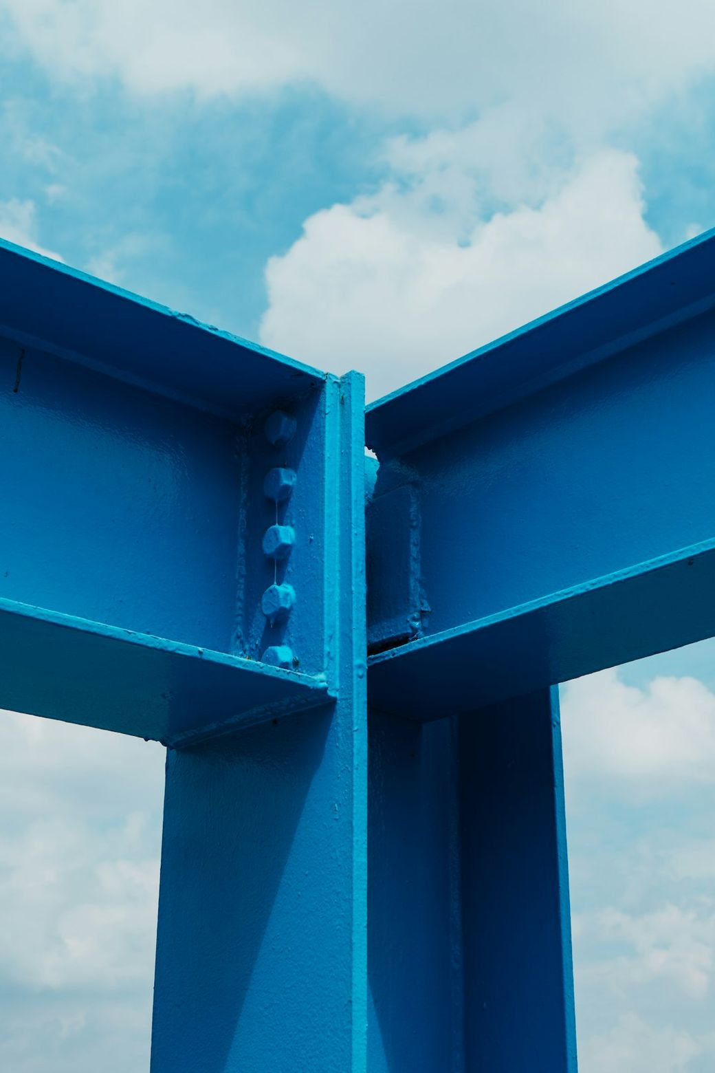 The image features a close-up view of a bright blue steel structure with two intersecting beams. The background includes a cloudy sky, adding contrast to the industrial elements. — Credit: Photo by Declan Sun on Unsplash