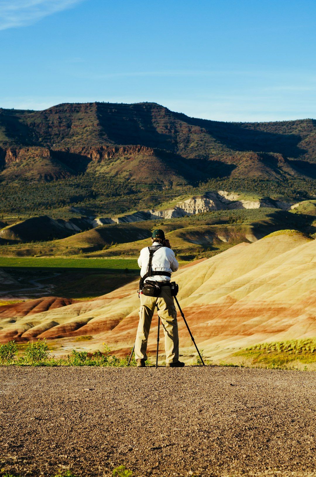 A photographer stands on a rocky outcrop capturing the scenic view of colorful, layered hills and mountainous terrain. The landscape features vibrant hues of red, orange, and yellow, indicating geological diversity.