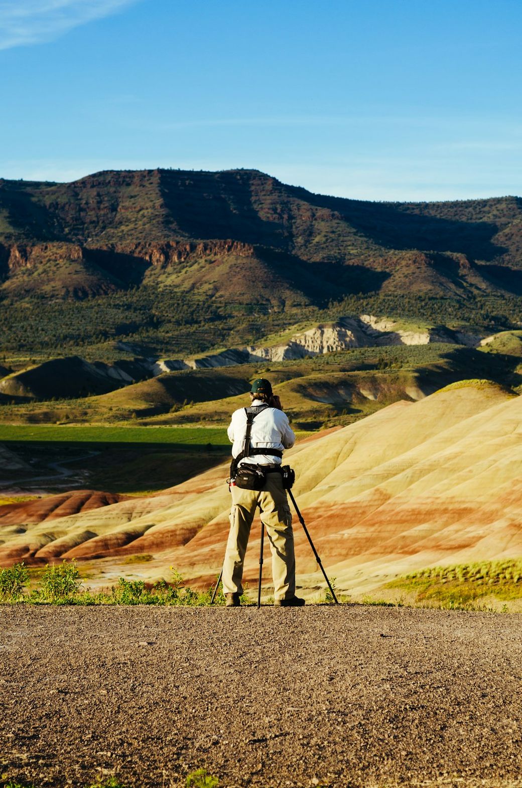 A photographer stands on a rocky outcrop capturing the scenic view of colorful, layered hills and mountainous terrain. The landscape features vibrant hues of red, orange, and yellow, indicating geological diversity. — Credit: Photo by Steven Weeks on Unsplash