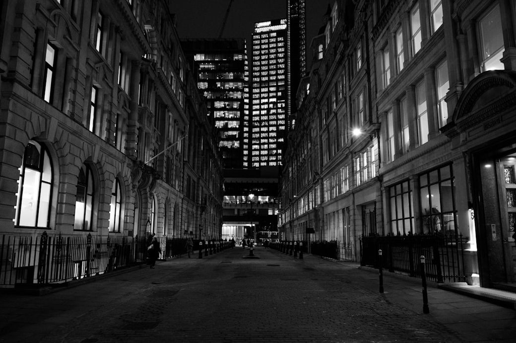 A narrow, cobblestone street at night is flanked by historical buildings with large windows illuminated, showcasing a contrast between the old architecture and the modern skyscraper in the background. The atmosphere is quiet and reflective, typical of a cityscape after dark. — Credit: Photo by Mario Mesaglio on Unsplash