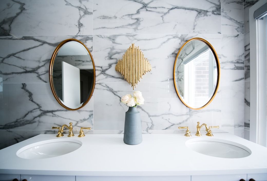 The image showcases a modern bathroom featuring dual golden faucets and oval mirrors set against a marble wall. A decorative wall piece and a simple flower arrangement add elegance to the space. — Credit: Photo by Christian Mackie on Unsplash
