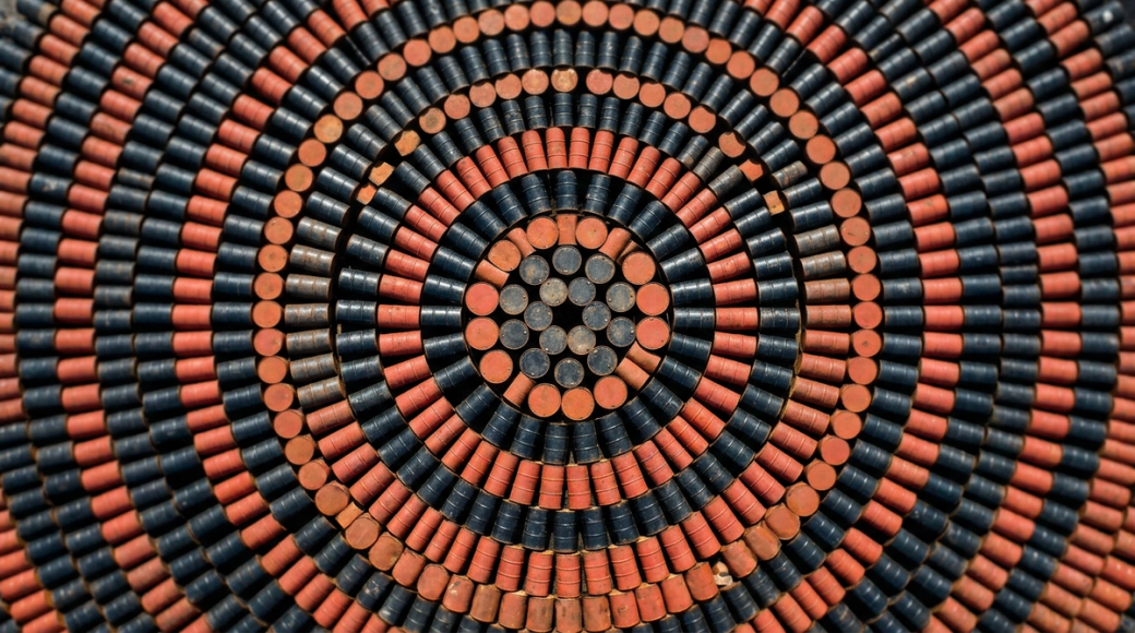 The image features an abstract aerial perspective of a vast storage yard filled with oil drums arranged in intricate concentric circles. The alternating black and rust-red barrels create a mesmerizing radial pattern, casting bright rims in the midday sunlight and forming deep shadows in the narrow gaps, suggesting a large-scale commodity storage environment.