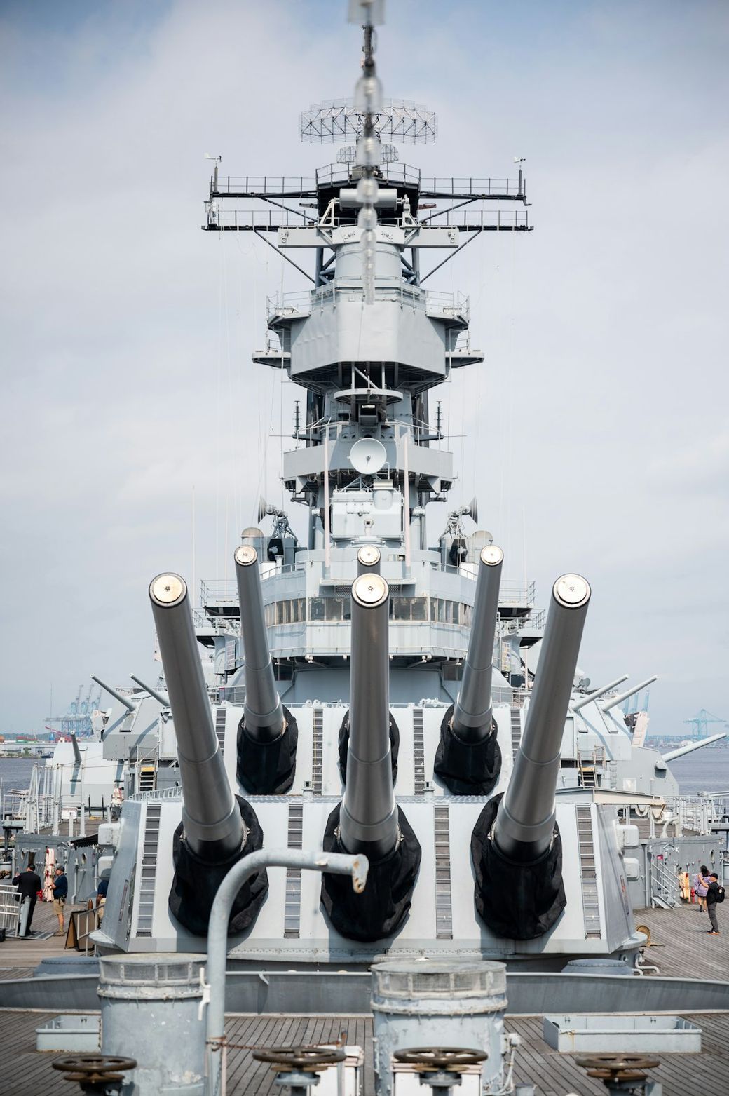 The image captures the bow of a battleship with multiple large naval guns prominently displayed. The setting is on the deck of the ship, with a cloudy sky in the background. — Credit: Photo by Peter Pryharski on Unsplash