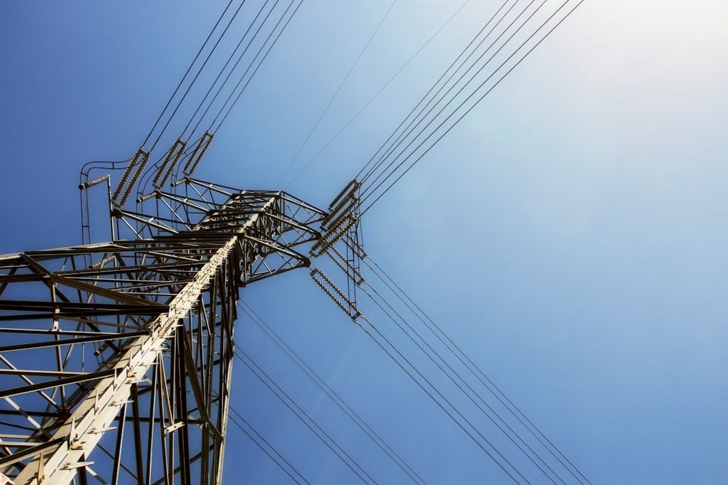 The image features a tall electrical transmission tower against a clear blue sky. The structure showcases the intricate networking of high-voltage power lines stretching into the distance. — Credit: Photo by Jason Richard on Unsplash