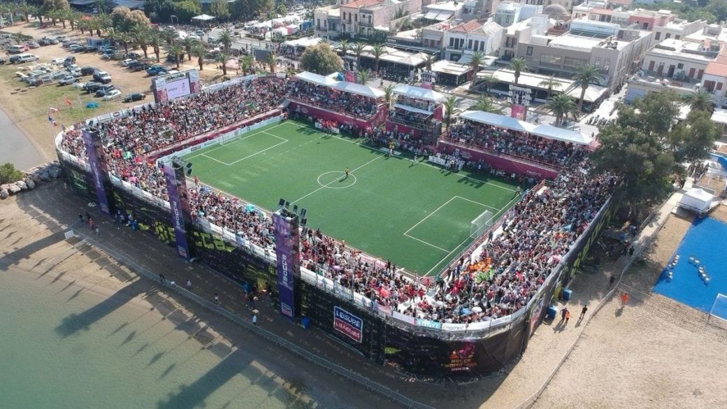 An aerial view of a crowded temporary soccer field set up near a beach, showcasing a vibrant atmosphere with fans gathered around. Various banners and signage can be seen, emphasizing the lively setting of this sporting event.