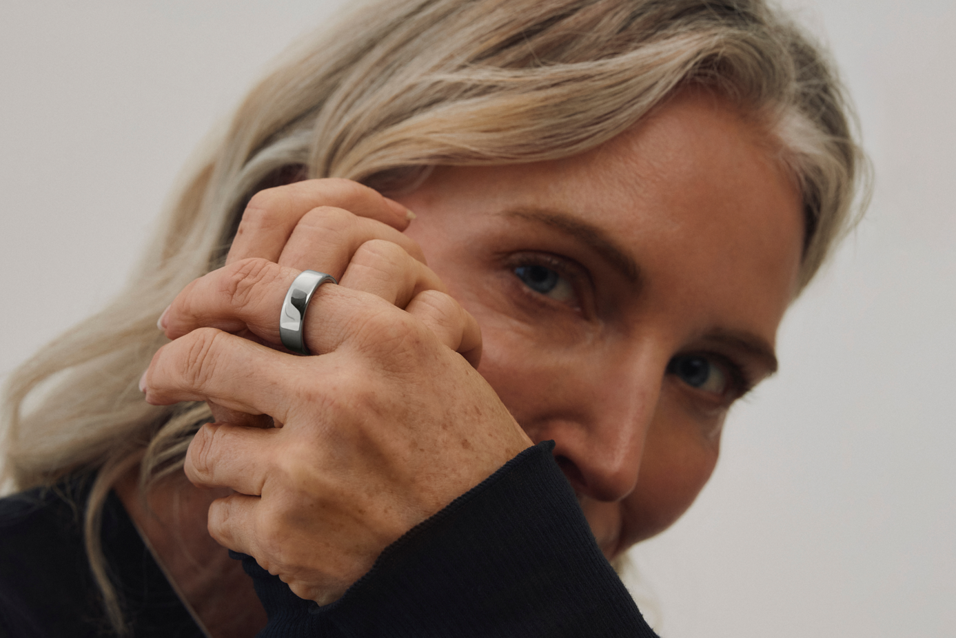 A close-up portrait of a woman with long, light-colored hair holding her hand near her face, showcasing a silver ring on her finger. The background is neutral, emphasizing the subject's features and the jewelry.