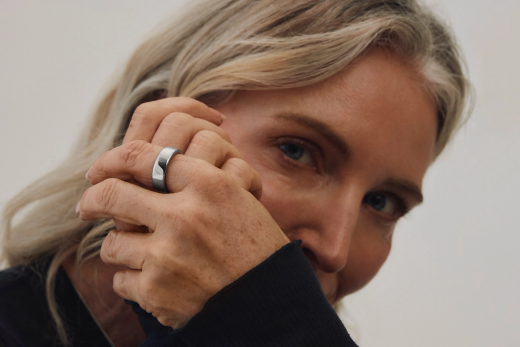 A close-up portrait of a woman with long, light-colored hair holding her hand near her face, showcasing a silver ring on her finger. The background is neutral, emphasizing the subject's features and the jewelry.