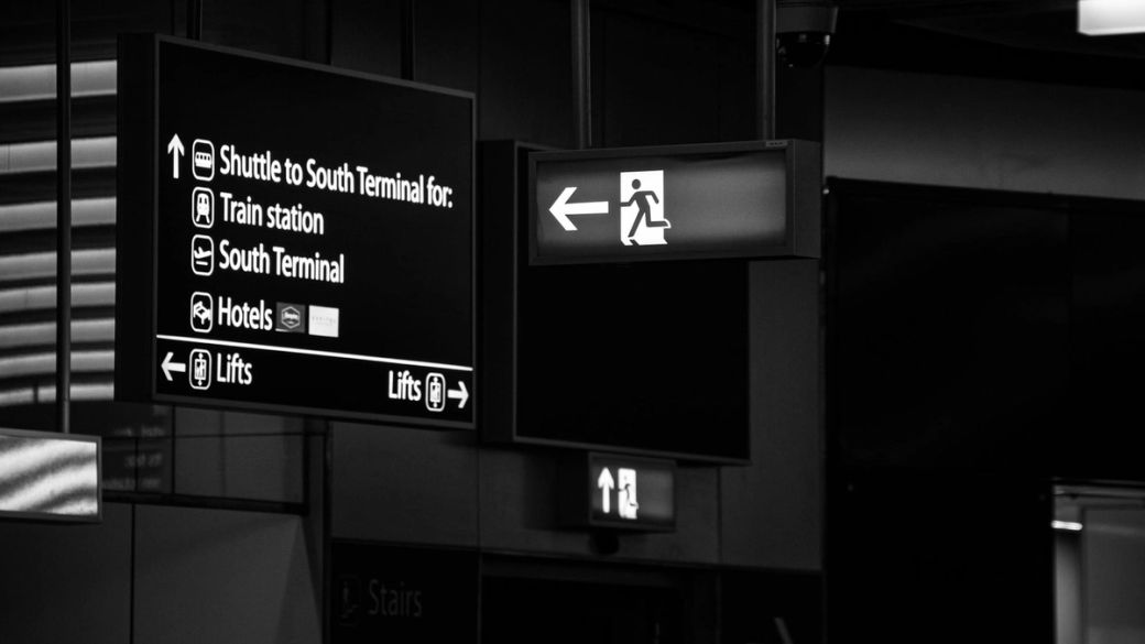 The image shows directional signage in an airport or transport hub, highlighting various services such as shuttle services, hotels, and lifts. The signs are in black and white, suggesting a modern and functional design.