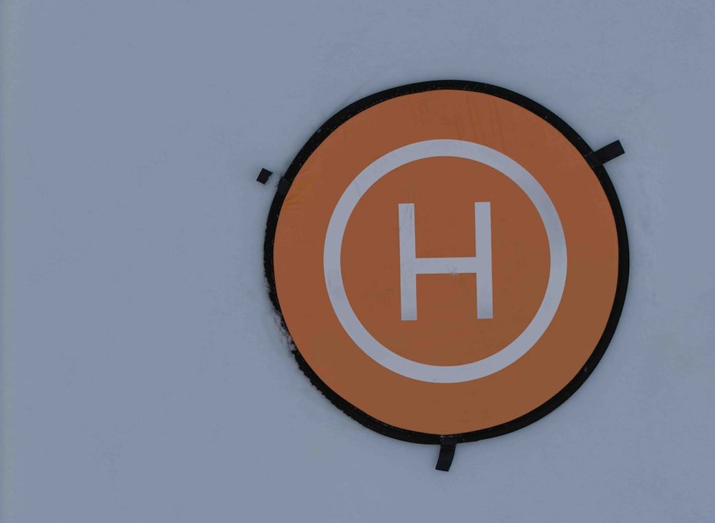 The image features a prominent orange circular sign with a white letter 'H' in the center, indicating a helipad. The sign is displayed against a light-colored background. — Credit: Photo by Bernd 📷 Dittrich on Unsplash