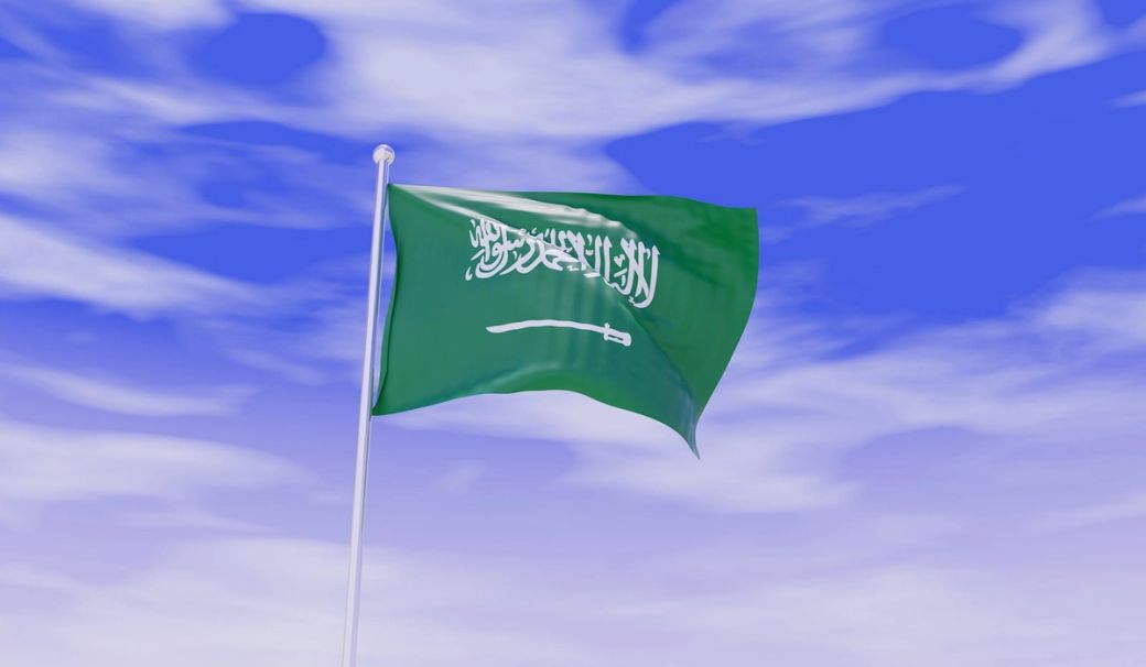 The image features the flag of Saudi Arabia fluttering against a blue sky. The flag prominently displays the Arabic inscription and a sword beneath it, symbolizing the nation’s identity and values. — Credit: Photo by Akhilesh Sharma / Unsplash