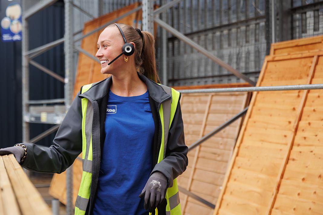 A woman wearing a headset and high-visibility vest smiles while working in a lumber yard. Wooden boards are stacked behind her, indicating a construction or retail setting.