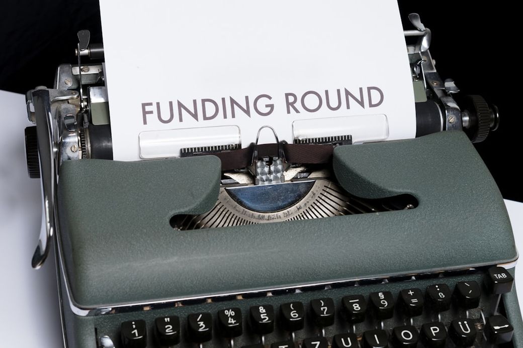 This image features a vintage typewriter with a sheet of paper inserted, displaying the text 'FUNDING ROUND'. The typewriter is positioned on a white surface, emphasizing its retro design. — Credit: Photo by Markus Winkler on Unsplash