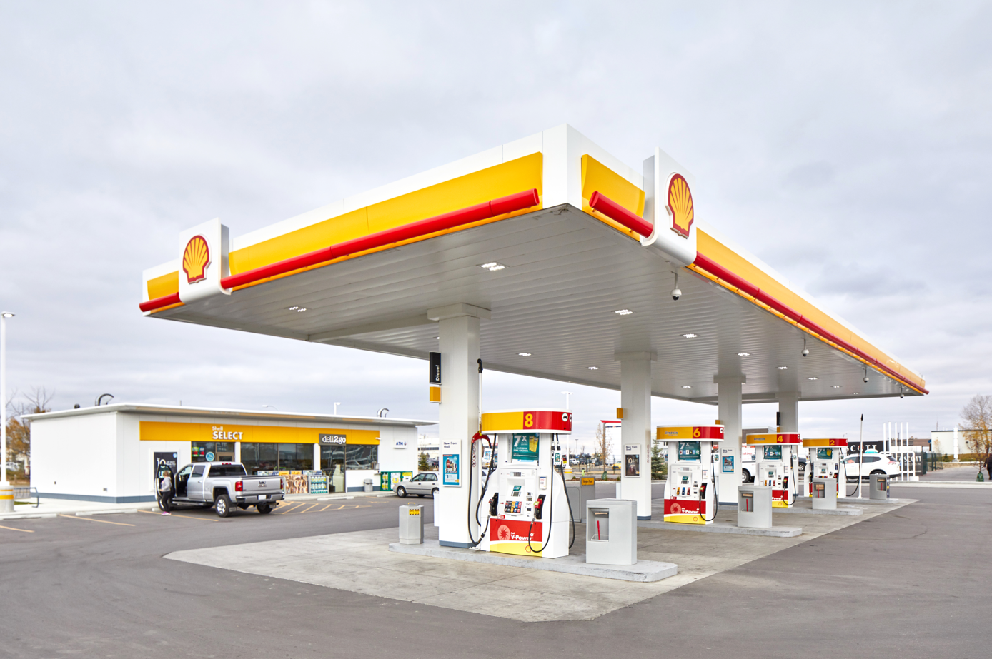 The image shows a Shell gas station with several fuel pumps under a covered area. There is a convenience store in the background, indicating a service station setup.