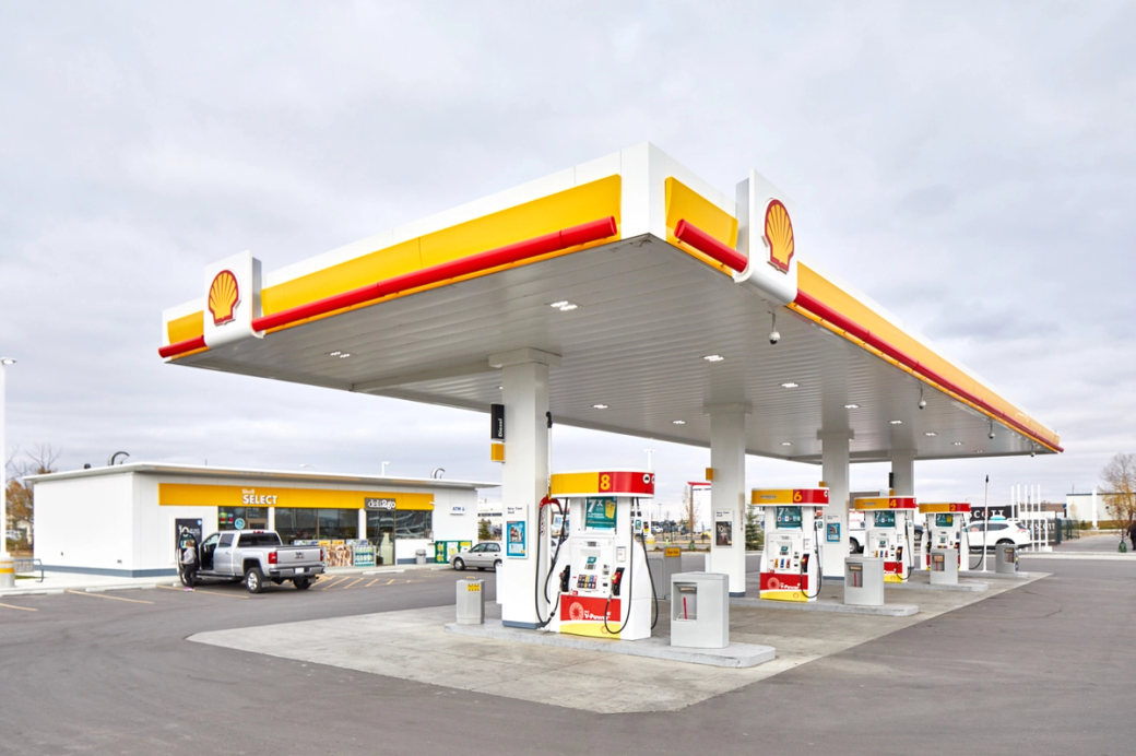 The image shows a Shell gas station with several fuel pumps under a covered area. There is a convenience store in the background, indicating a service station setup.