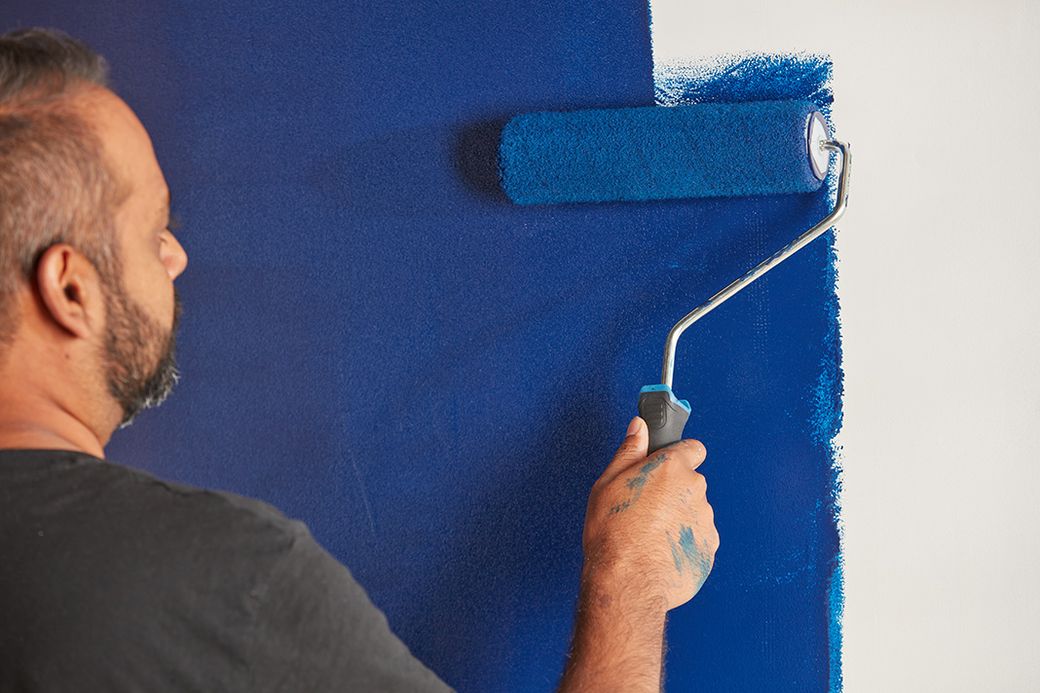 A man is painting a wall with a blue paint roller. The image focuses on the man applying paint, showcasing the action of home improvement or renovation.