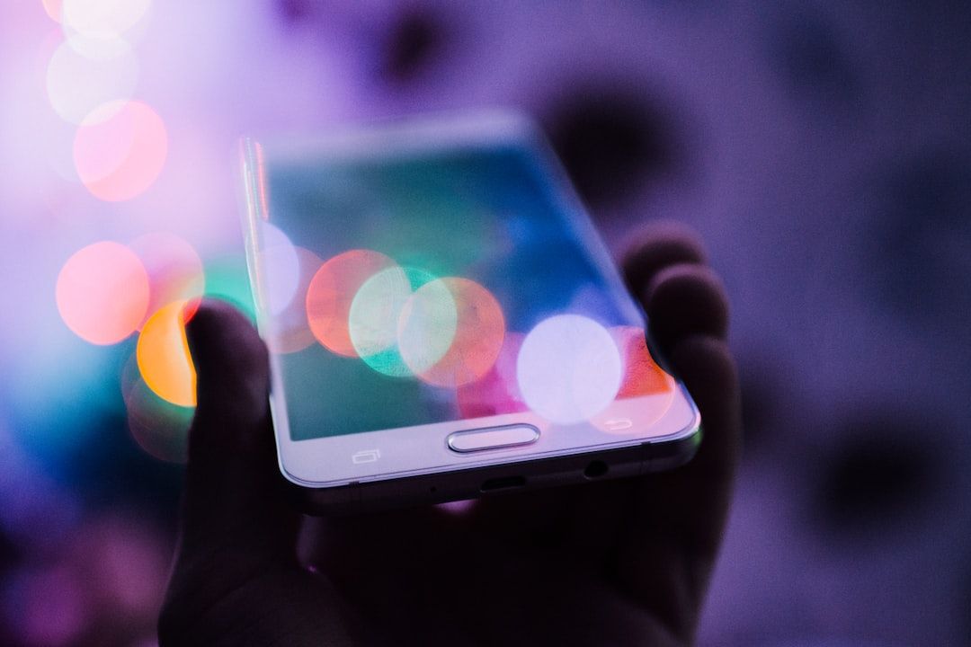 A hand holding a smartphone with a blurred, colorful background featuring bokeh lights. The image captures a modern, tech-savvy setting reminiscent of nightlife or creative digital environments.