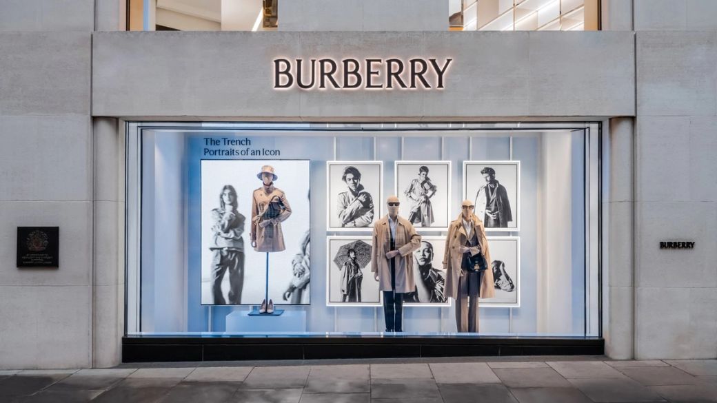 The image depicts a Burberry retail storefront showcasing a fashion display. The window features mannequins dressed in trench coats along with a promotional graphic titled 'The Trench Portraits of an Icon'.