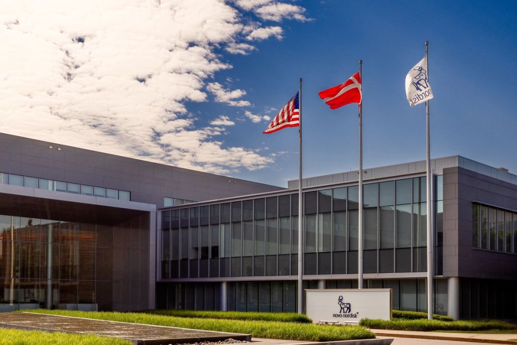 The image shows a modern corporate building featuring large glass windows and a clean architectural design. Three flags, including the American flag, are prominently displayed in front of the structure, suggesting a multinational presence.