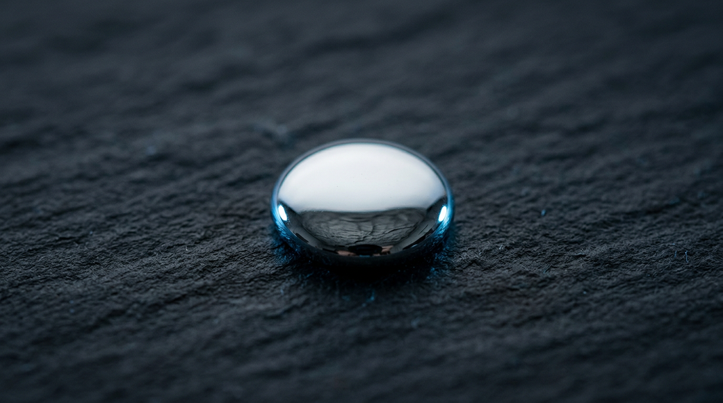 The image features a single droplet of liquid mercury perched on a rough, dark slab of slate. The droplet exhibits a smooth, mirror-like surface that reflects the texture of the slate beneath it, enhanced by a cool silver-white metallic tone and faint blue ambient light along its edge.